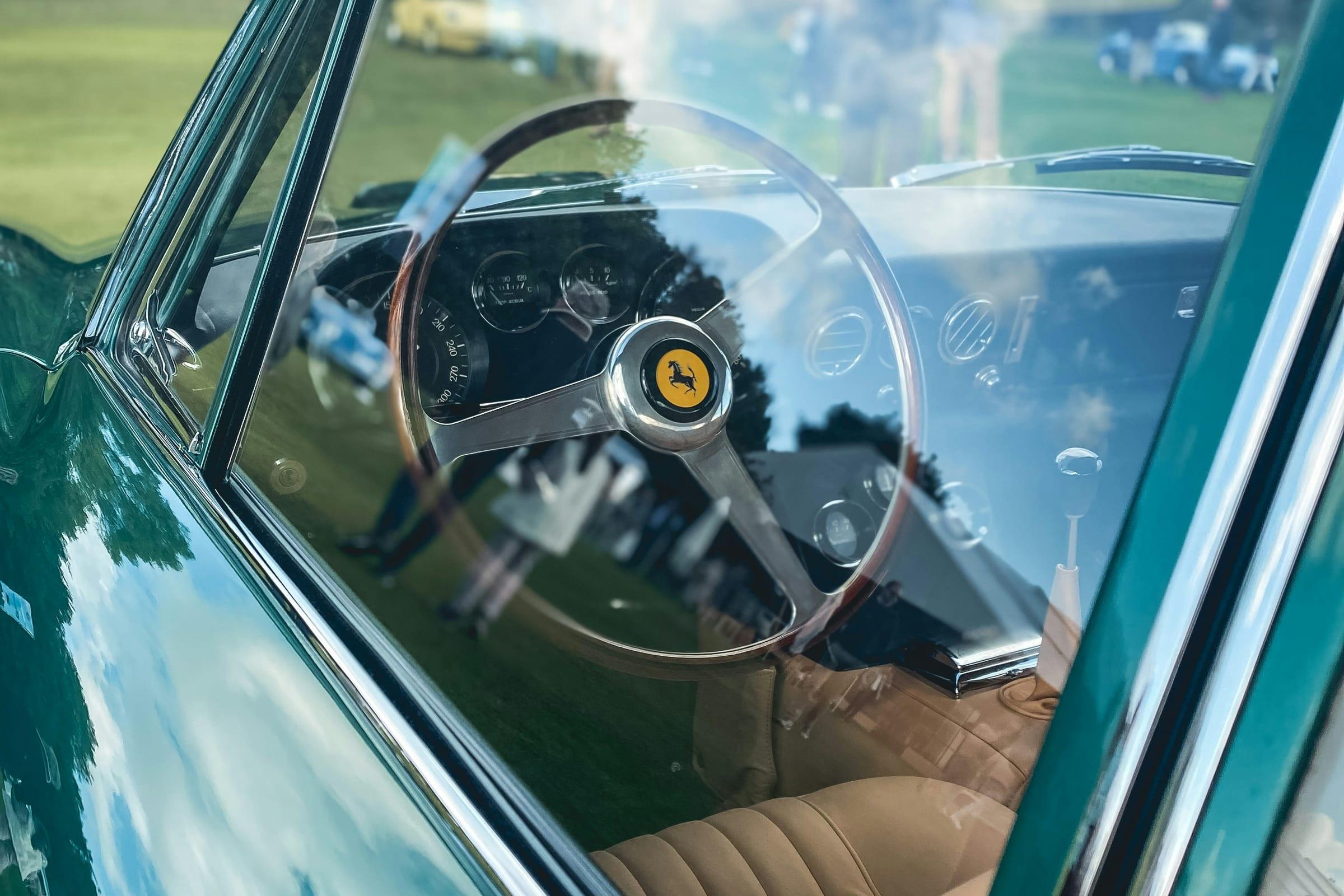 The cockpit of a classic sports car seen through the side glass, wood-rimmed wheel in shadow.