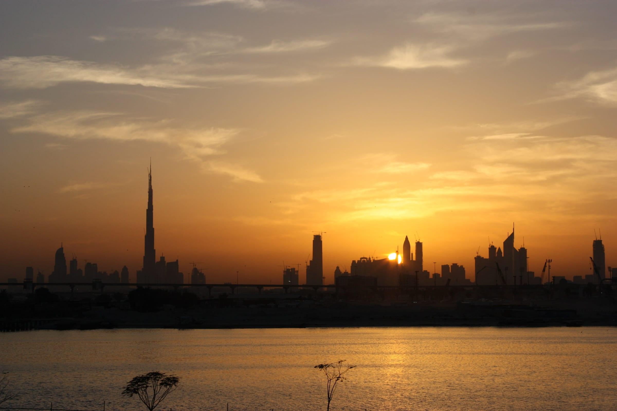 The Dubai skyline in silhouette at sunset, seen across open ground.